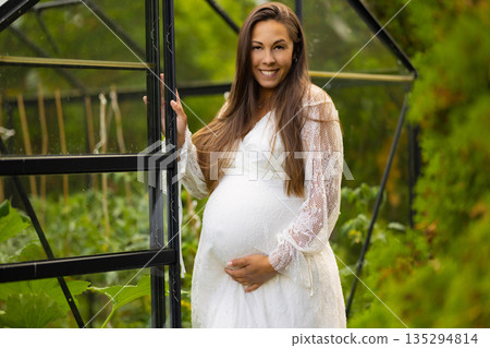 Pregnant woman smiling near a greenhouse surrounded by lush green foliage Pregnant woman smiling near a greenhouse surrounded by lush green foliage 135294814