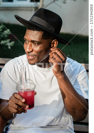 Man holding red drink outdoors in sunlight. Refreshment, relaxation, and summer lifestyle through casual beverage moment Man holding red drink outdoors in sunlight. Refreshment, relaxation, and summer lifestyle through casual beverage moment 135294869