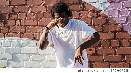 Man dancing with headphones against painted brick wall. Music enjoyment, personal freedom, and self expression through urban street culture Man dancing with headphones against painted brick wall. Music enjoyment, personal freedom, and self expression through urban street culture 135294876