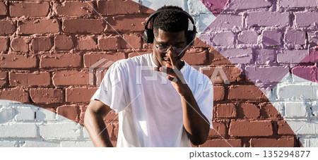 Man dancing happily with headphones by colorful wall. Positive mood, music therapy, and carefree lifestyle through urban street performance Man dancing happily with headphones by colorful wall. Positive mood, music therapy, and carefree lifestyle through urban street performance 135294877