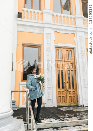 African american Woman holding bouquet of white roses in winter sunlight outdoors. Joy, tenderness, and individuality expressed through urban winter lifestyle and seasonal atmosphere. African american Woman holding bouquet of white roses in winter sunlight outdoors. Joy, tenderness, and individuality expressed through urban winter lifestyle and seasonal atmosphere. 135295381