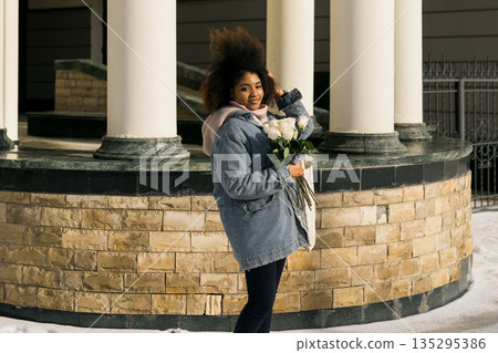 African american Woman holding bouquet of white roses in winter sunlight outdoors. Joy, tenderness, and individuality expressed through urban winter lifestyle and seasonal atmosphere. 135295386
