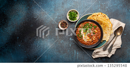 Traditional Indian Punjabi dish Dal makhani with lentils and beans in black bowl served with naan flat bread, fresh cilantro and two spoons on blue concrete rustic table top view. Space for text 135295548