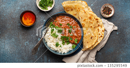 Traditional Indian Punjabi dish Dal makhani with lentils and beans in black bowl served with basmati rice, naan flat bread, fresh cilantro and spoon on blue concrete rustic table top view 135295549