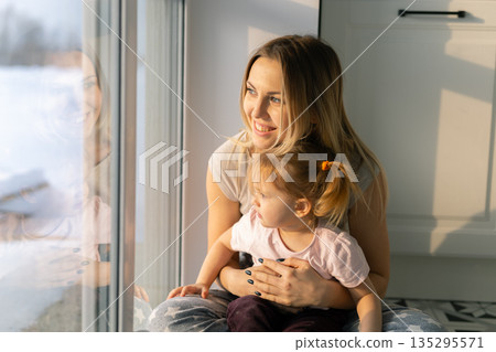 Mother holding her daughter close while watching through the window in soft sunlight. Family love, emotional peace, and gentle connection in quiet home atmosphere. Mother holding her daughter close while watching through the window in soft sunlight. Family love, emotional peace, and gentle connection in quiet home atmosphere. 135295571