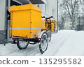 Traditional german yellow cargo postal mail bicycle parked at residential building mailbox at cold snowy winter snowfall day. Urban delivery service daily logistics work. Snow street cold weather 135295582