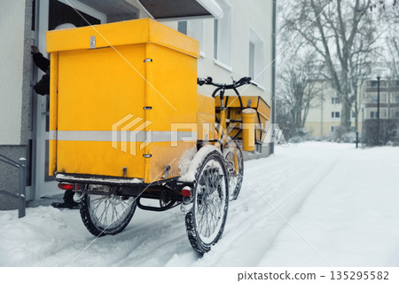 Traditional german yellow cargo postal mail bicycle parked at residential building mailbox at cold snowy winter snowfall day. Urban delivery service daily logistics work. Snow street cold weather 135295582