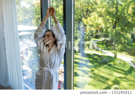 Pretty young woman in bathrobe standing near the window 135295811