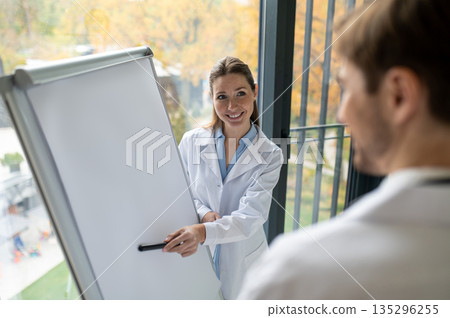 Female doctor standing near the flipchart and presenting something to a new doctor Female doctor standing near the flipchart and presenting something to a new doctor 135296255