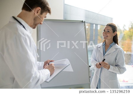 Female doctor standing near the flipchart and presenting something to a new doctor 135296259