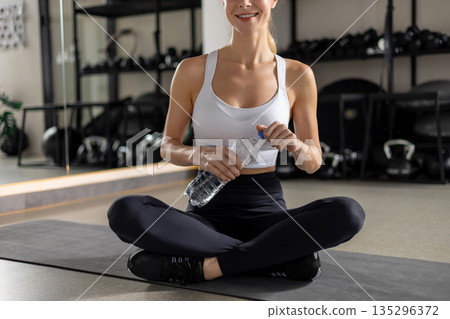 Young woman sitting on the mat with a bottle of water in hands Young woman sitting on the mat with a bottle of water in hands 135296372