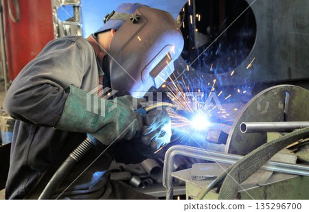 Close-up of a worker with protective mask and gloves welding metal parts in a workshop. Bright sparks and blue light during the industrial manufacturing process. Close-up of a worker with protective mask and gloves welding metal parts in a workshop. Bright sparks and blue light during the industrial manufacturing process. 135296700