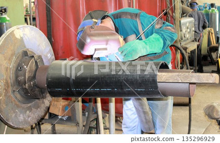 Industrial worker with protective helmet and green gloves performing TIG welding on a large black metal pipe. Skilled labor in a mechanical engineering factory. Industrial worker with protective helmet and green gloves performing TIG welding on a large black metal pipe. Skilled labor in a mechanical engineering factory. 135296929