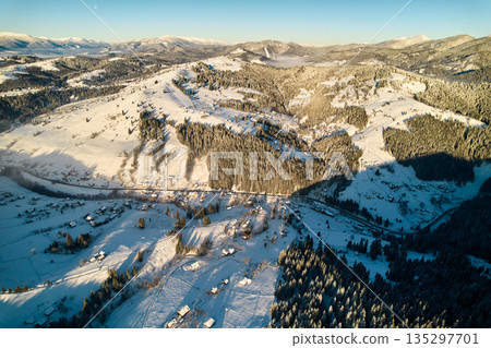 Aerial view of snow-covered valley with winding road passing through small village. Landscape features gently rolling hills, dense forests, and distant mountains under clear blue sky, with soft mist. 135297701
