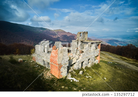 Ancient, crumbling stone building atop grassy hill, surrounded by vast landscape of rolling mountains and dense forests. Clear sky with scattered clouds adds depth to panoramic view. 135297712