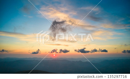 Sun sets behind rolling hills, casting warm, golden glow across sky. Wispy clouds, tinged with hues of orange and yellow, stretch across deepening blue sky. Serene and expansive landscape. Sun sets behind rolling hills, casting warm, golden glow across sky. Wispy clouds, tinged with hues of orange and yellow, stretch across deepening blue sky. Serene and expansive landscape. 135297716