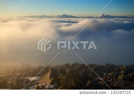 Sunrise over snow-covered mountain peak. Warm golden light of rising sun bathes clouds in soft hues of orange and pink. Snow and scattered rocks in foreground add texture and depth to scene. 135297725