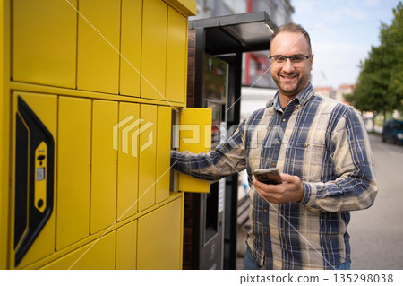 Man using smartphone near yellow delivery lockers in an urban area during daylight 135298038