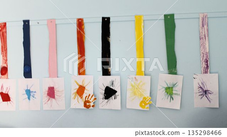 A row of creative handmade artworks by preschool children displayed on a light blue classroom wall. The crafts include colorful painted strips and finger-painted handprints on white paper, showcasing A row of creative handmade artworks by preschool children displayed on a light blue classroom wall. The crafts include colorful painted strips and finger-painted handprints on white paper, showcasing 135298466