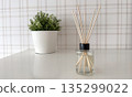 A minimalist home decor composition featuring a clear glass reed diffuser bottle with wooden sticks standing on a glossy white table 135299022