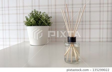 A minimalist home decor composition featuring a clear glass reed diffuser bottle with wooden sticks standing on a glossy white table A minimalist home decor composition featuring a clear glass reed diffuser bottle with wooden sticks standing on a glossy white table 135299022