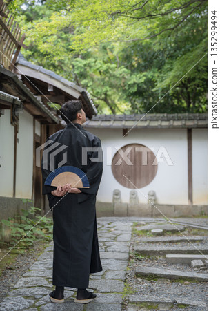 A man in a black kimono standing on a stone pavement, holding a folding fan. Japan 135299494