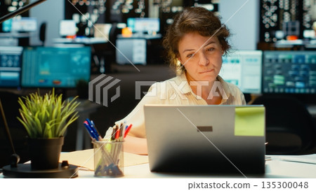 Businesswoman stretching her upper back and neck muscles, correcting the posture and neck stiffness for relief and relaxation. Intense office activity due to overtime late at night. Camera B. Businesswoman stretching her upper back and neck muscles, correcting the posture and neck stiffness for relief and relaxation. Intense office activity due to overtime late at night. Camera B. 135300048