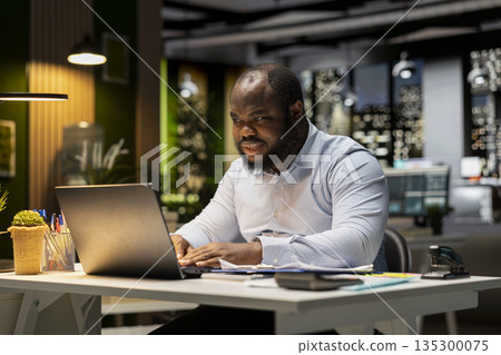 Black business consultant working late at night on proofreading documents, writing important reports on his laptop. Creating a summary and designing strategy to align with company objectives. Black business consultant working late at night on proofreading documents, writing important reports on his laptop. Creating a summary and designing strategy to align with company objectives. 135300075