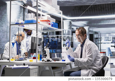 Male researcher mixing matter in a flask with pipette for research study, biotechnology research for laboratory testing. Molecular biology and medical research through advanced preparation. Male researcher mixing matter in a flask with pipette for research study, biotechnology research for laboratory testing. Molecular biology and medical research through advanced preparation. 135300079