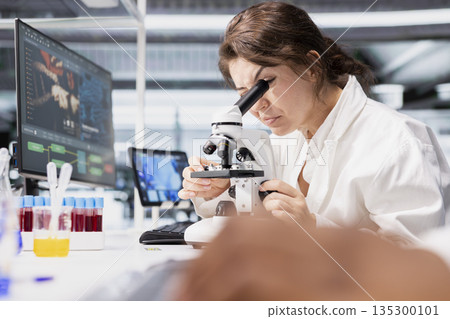 Close up of histology technician in lab using scientific microscope for cellular analysis. Woman studies sample inside modern research facility with optical device equipment Close up of histology technician in lab using scientific microscope for cellular analysis. Woman studies sample inside modern research facility with optical device equipment 135300101