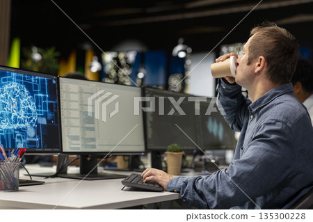 Developer drinking coffee in office, reviewing AI system metrics, monitoring performance dashboards. Tech company IT expert enjoying caffeinated beverage to wake up while using program on computer 135300228