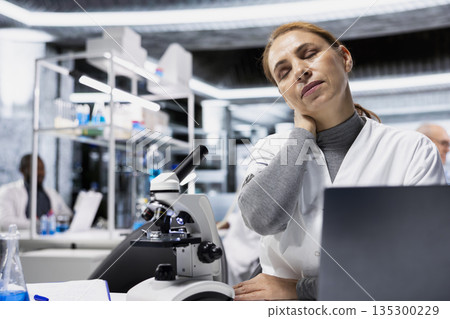 Exhausted scientist in lab suffering neck ache while documenting clinical trial outcomes on laptop. Fatigued researcher massaging stiff muscles in lab while updating database system 135300229