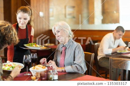 In cafe, girl waitress serves catering establishments mature couple visitor, puts food on table. In cafe, girl waitress serves catering establishments mature couple visitor, puts food on table. 135303317