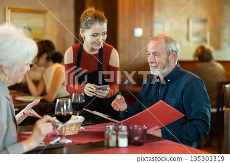 At cafe, girl waitress communicates with mature couple of customer, accepts order 135303319