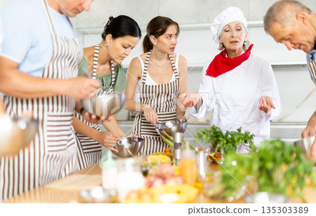 Adult students prepare food according to recipe in cooking class led by female chef 135303389