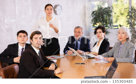 Young woman turning on presentation on projector while office coworkers of different ages making notes at table with laptops Young woman turning on presentation on projector while office coworkers of different ages making notes at table with laptops 135303485