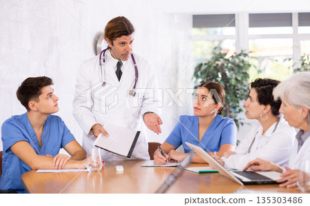 Male chief doctor discussing reports from colleagues of different ages in conference room Male chief doctor discussing reports from colleagues of different ages in conference room 135303486