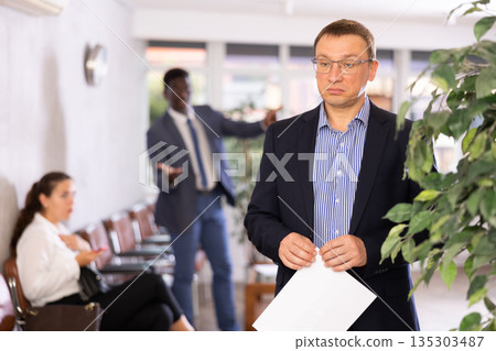 Upset middle-aged man standing in waiting room with his back to person who is shouting at him Upset middle-aged man standing in waiting room with his back to person who is shouting at him 135303487