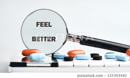 Pills are placed on a white surface, and a magnifying glass with the words FEEL BETTER is resting on them. The concept represents medical care and improved health 135303492