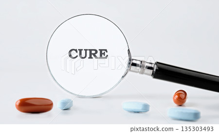 The word Cure is magnified by a magnifying glass resting near pills of different sizes and colors scattered on a clean, white backdrop in a studio setting 135303493