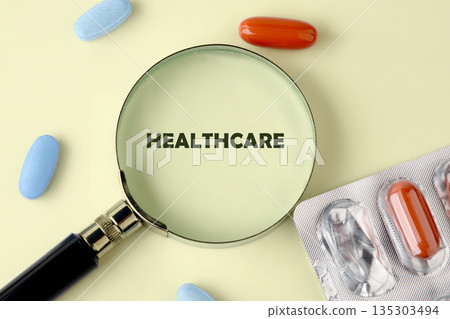 Magnifying glass shows the word Healthcare. Pills, including orange capsules in blister packs and individual blue pills, surround the magnifying glass on a light yellow surface 135303494