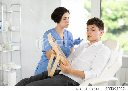 Man sits in chair at a doctor appointment with beautician. Woman doing facial examination before surgery 135303622