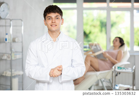 Young man doctor posing in doctor's cabinet with patient lying on clinical chair Young man doctor posing in doctor's cabinet with patient lying on clinical chair 135303632