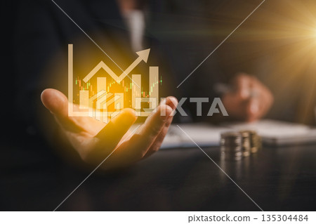 Business finance growth concept with hand holding holographic chart, rising arrow and candlestick data, coins on desk, showing investment performance, profit and market analysis strategy. Business finance growth concept with hand holding holographic chart, rising arrow and candlestick data, coins on desk, showing investment performance, profit and market analysis strategy. 135304484