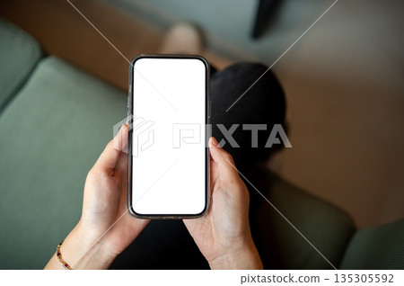Close up of woman's hands holding white screen smartphone while sitting cross legged on green sofa. 135305592