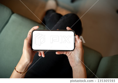 Close up of woman's hands holding white screen phone playing or watching while sitting on green sofa Close up of woman's hands holding white screen phone playing or watching while sitting on green sofa 135305593