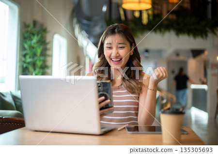 Asian woman holding phone raising a fist in happy or excited over laptop on table in cafe or canteen 135305693