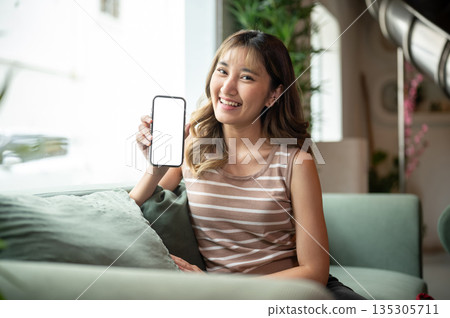 Asian woman showing white screen phone and sitting leaning against green sofa in a cafe or lounge. Asian woman showing white screen phone and sitting leaning against green sofa in a cafe or lounge. 135305711