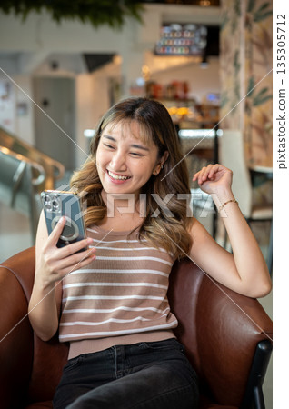 Pretty asian woman smiling as looking at smartphone while sitting on brown sofa in cafe or canteen. Pretty asian woman smiling as looking at smartphone while sitting on brown sofa in cafe or canteen. 135305712