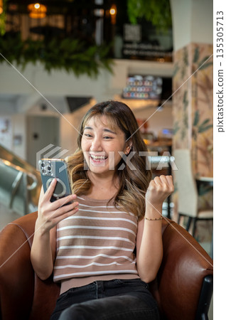 Asian woman making a fist as looking at smartphone while sitting on brown sofa in cafe or canteen. Asian woman making a fist as looking at smartphone while sitting on brown sofa in cafe or canteen. 135305713